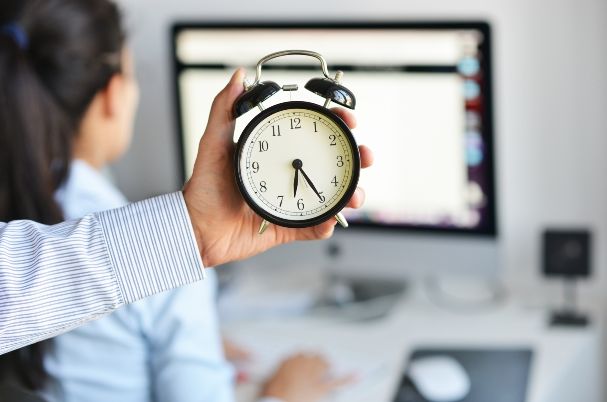 Overtime exemption photo concept shows a businessman's hand holding a clock with a female worker in the background who is typing at her computer.